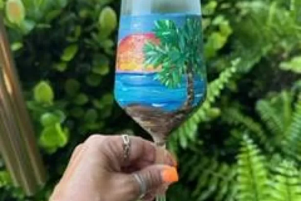 a hand with bright orange finger nails holding up a brightly painted wine glass with a beach scene on it
