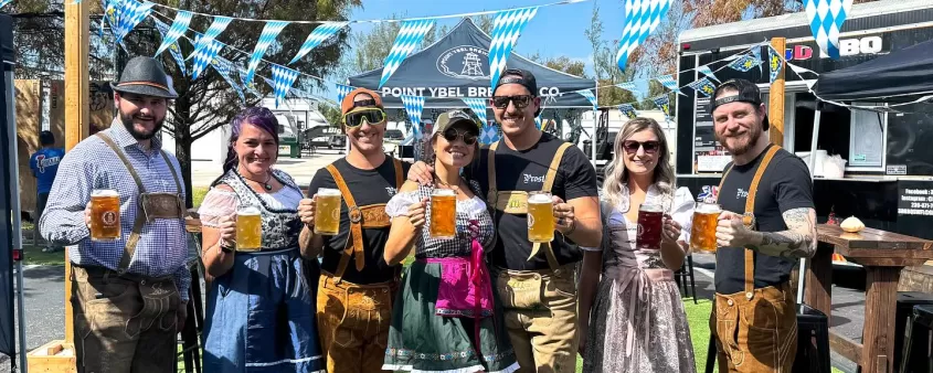 Group of people dressed in traditional Bavarian-style outfits holding large beer steins at an outdoor Oktoberfest-style event.